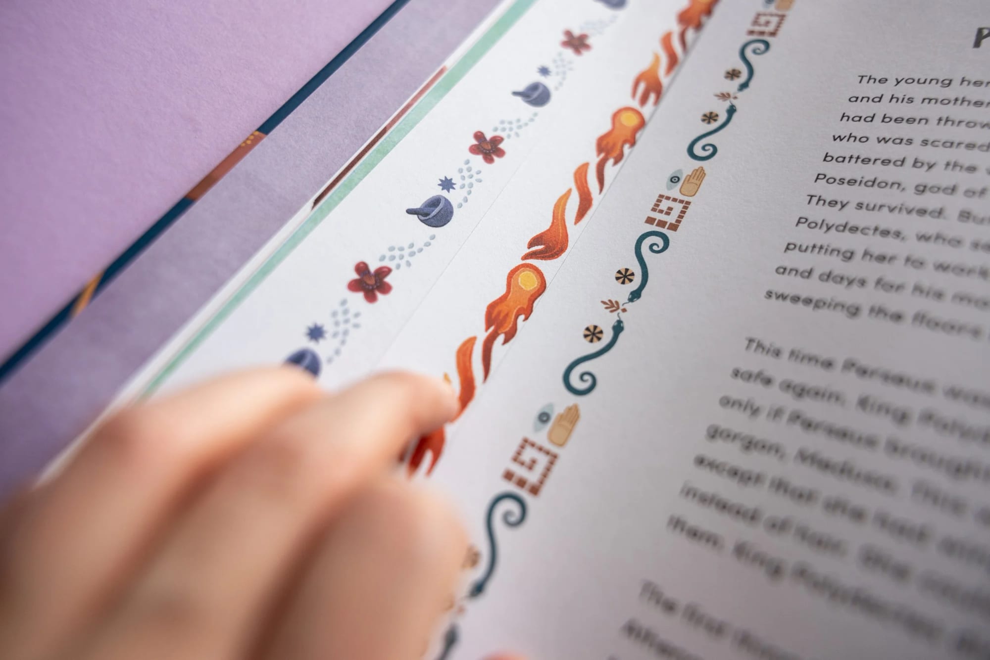 Close-up of a hand pointing at colorful decorative border on a printed page, with part of the text visible. The border features illustrations of flowers, flames, and abstract shapes.