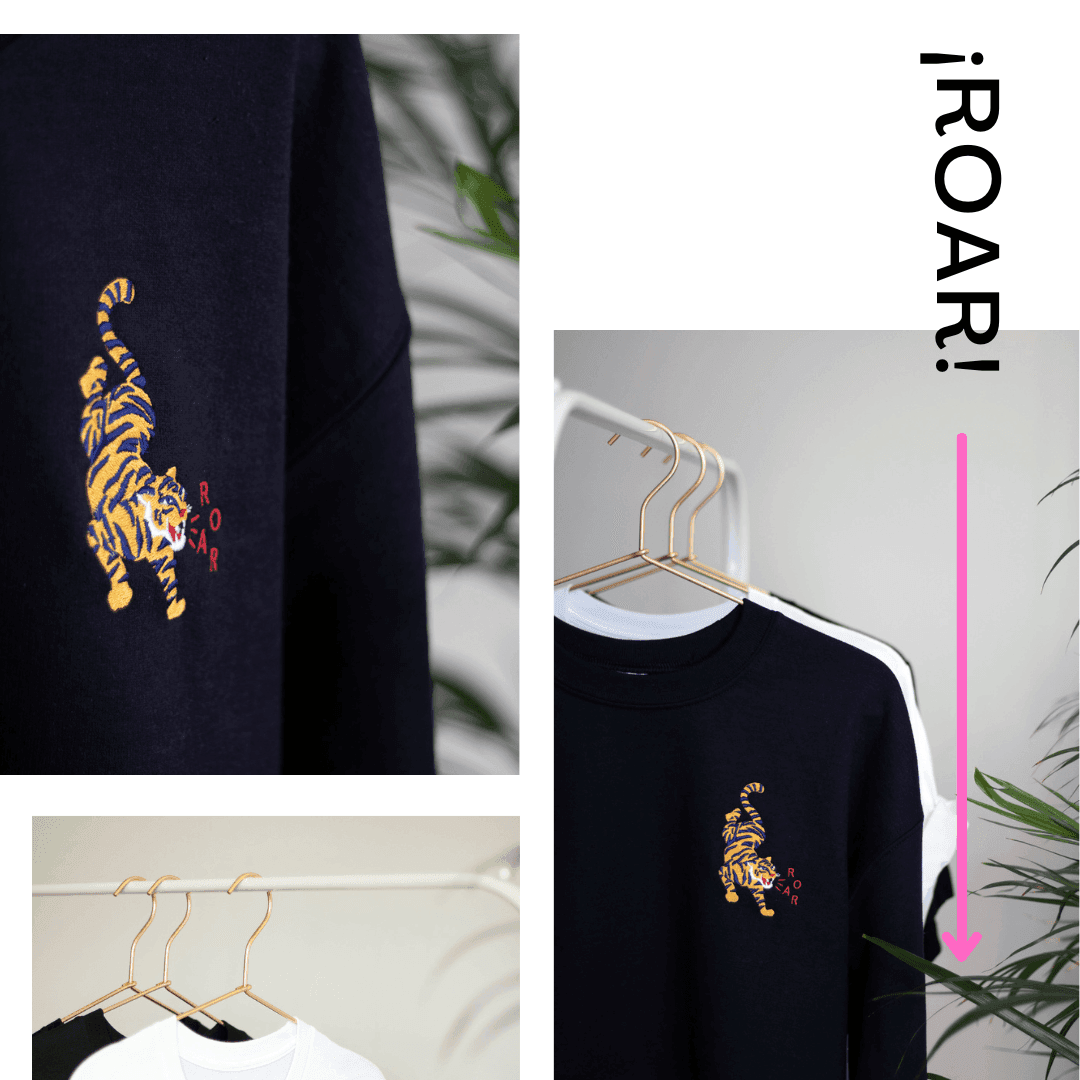 Close-up of a navy sweatshirt with an embroidered yellow and black tiger and the word 'ROAR' on a hanger, with other sweatshirts hanging nearby and green plants in the background.