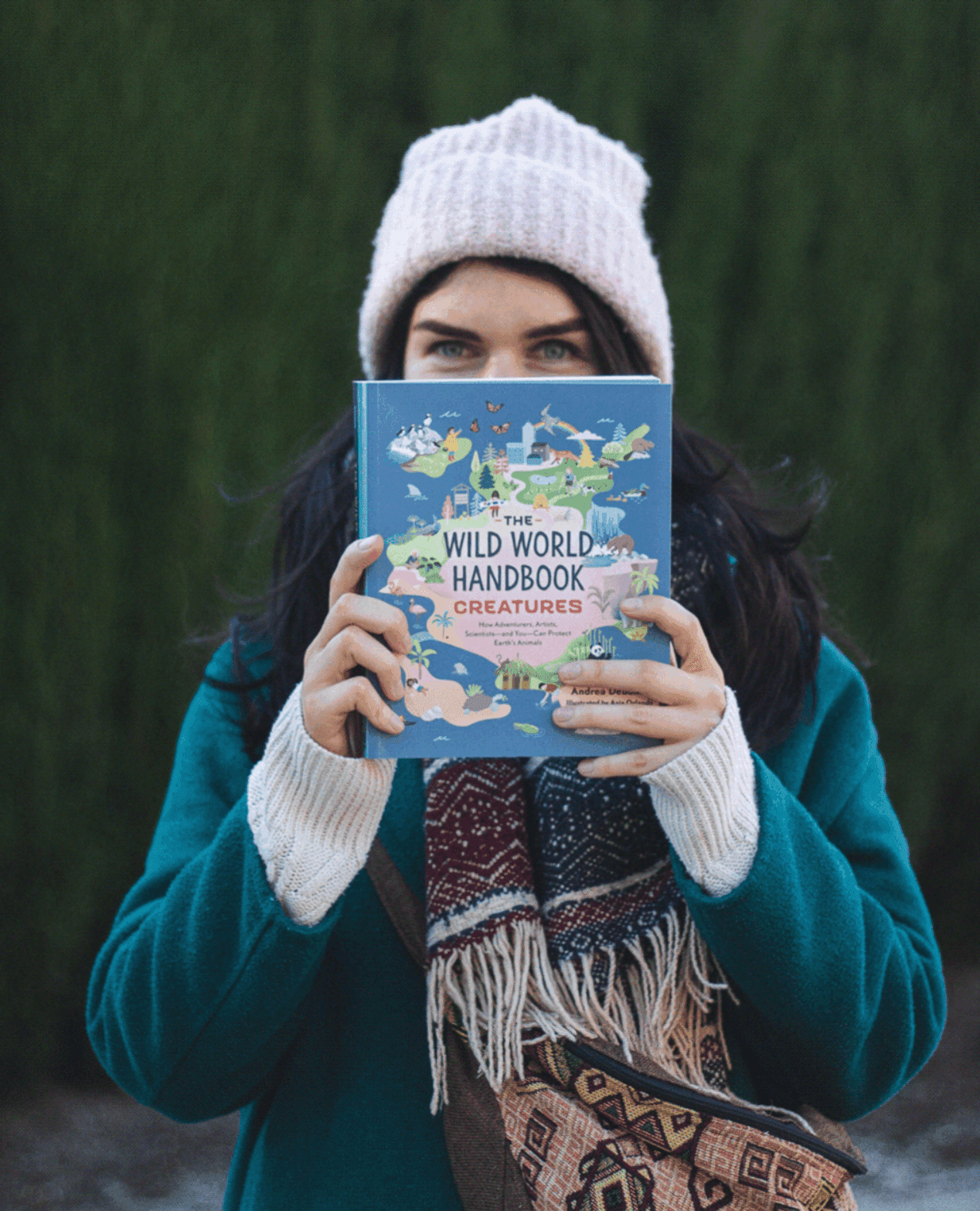 A woman wearing a pink knit hat and a teal coat holding a book titled 'The Wild World Handbook: Creatures' in front of her face, standing outdoors with trees in the background.