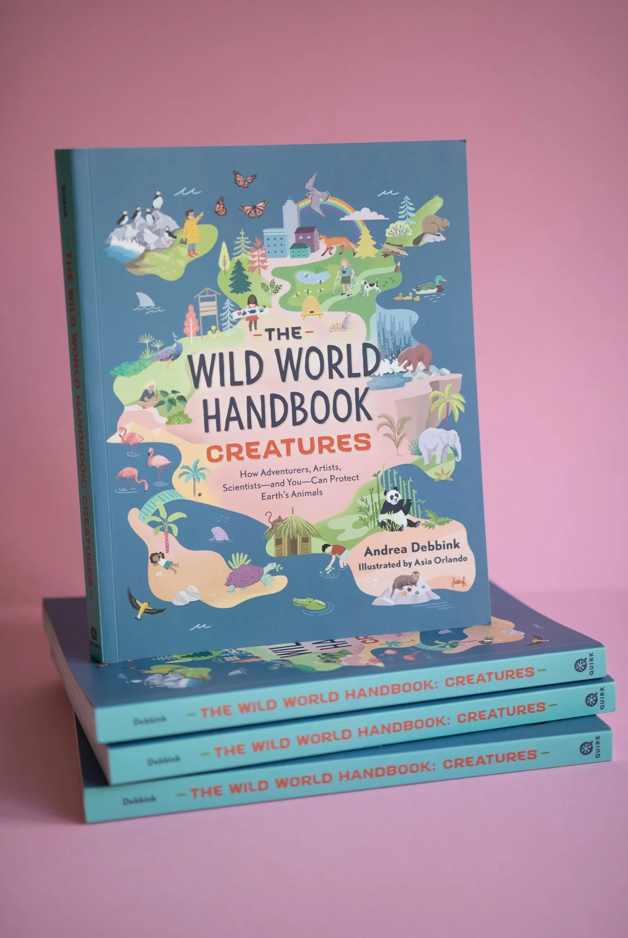 A colorful children's hardcover book titled "The Wild World Handbook: Creatures" is standing upright on a pink surface, with additional copies stacked in front of it. The cover features illustrations of various animals and nature scenes.