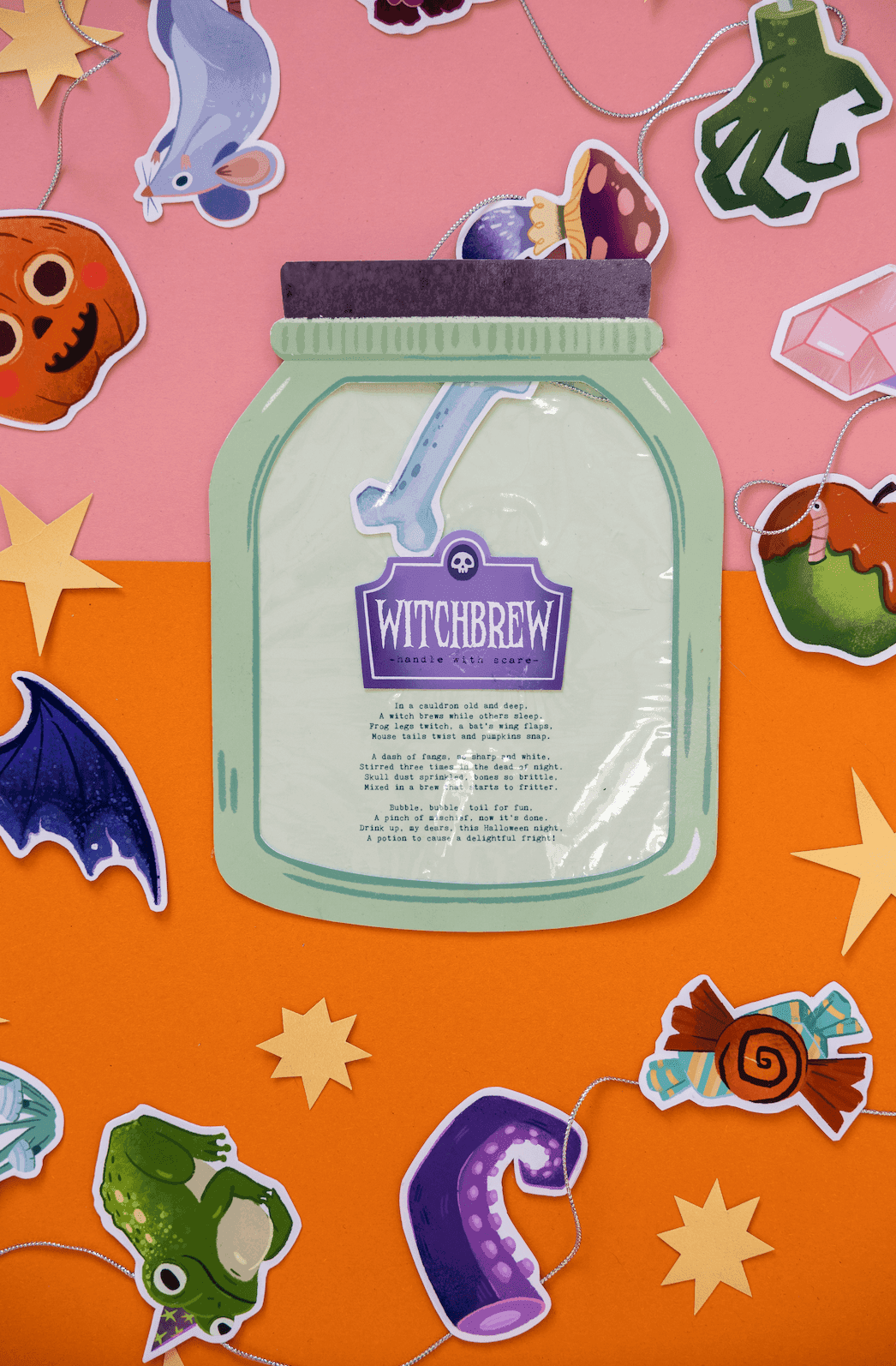 A Halloween themed display with a jar-shaped decoration labeled 'Witchbrew' and assorted Halloween stickers of a pumpkin, bat, lizard, spider, candy, moon, and stars surrounding it, with an orange and pink background.