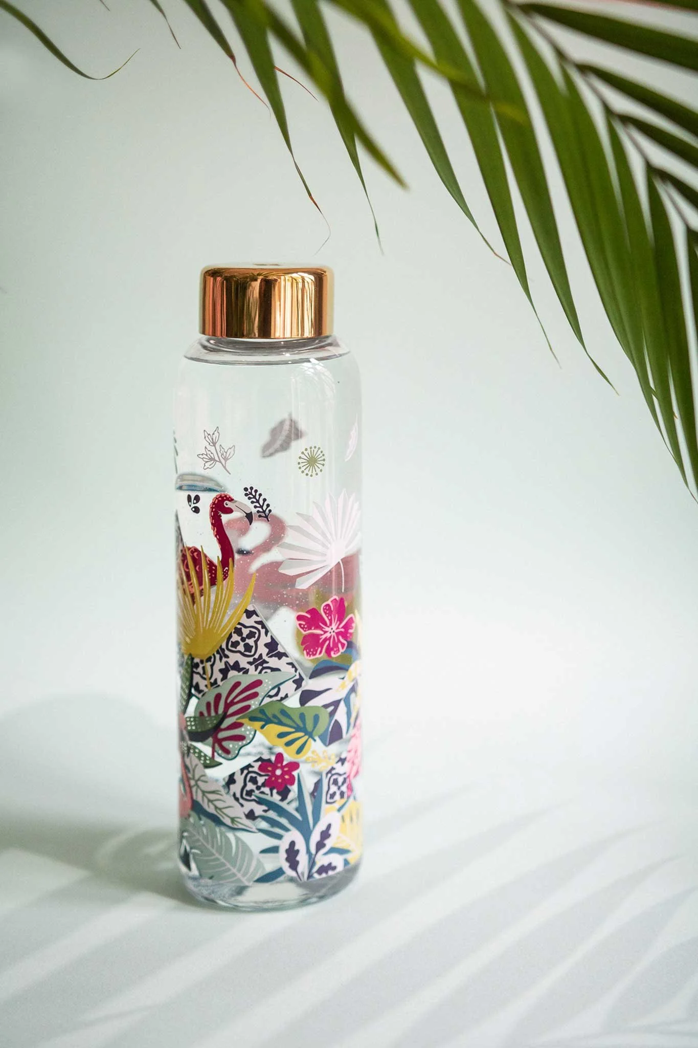 A clear water bottle with a gold cap, decorated with colorful tropical illustrations, including a pink flamingo, various leaves, and flowers, placed against a plain background near green palm leaves.