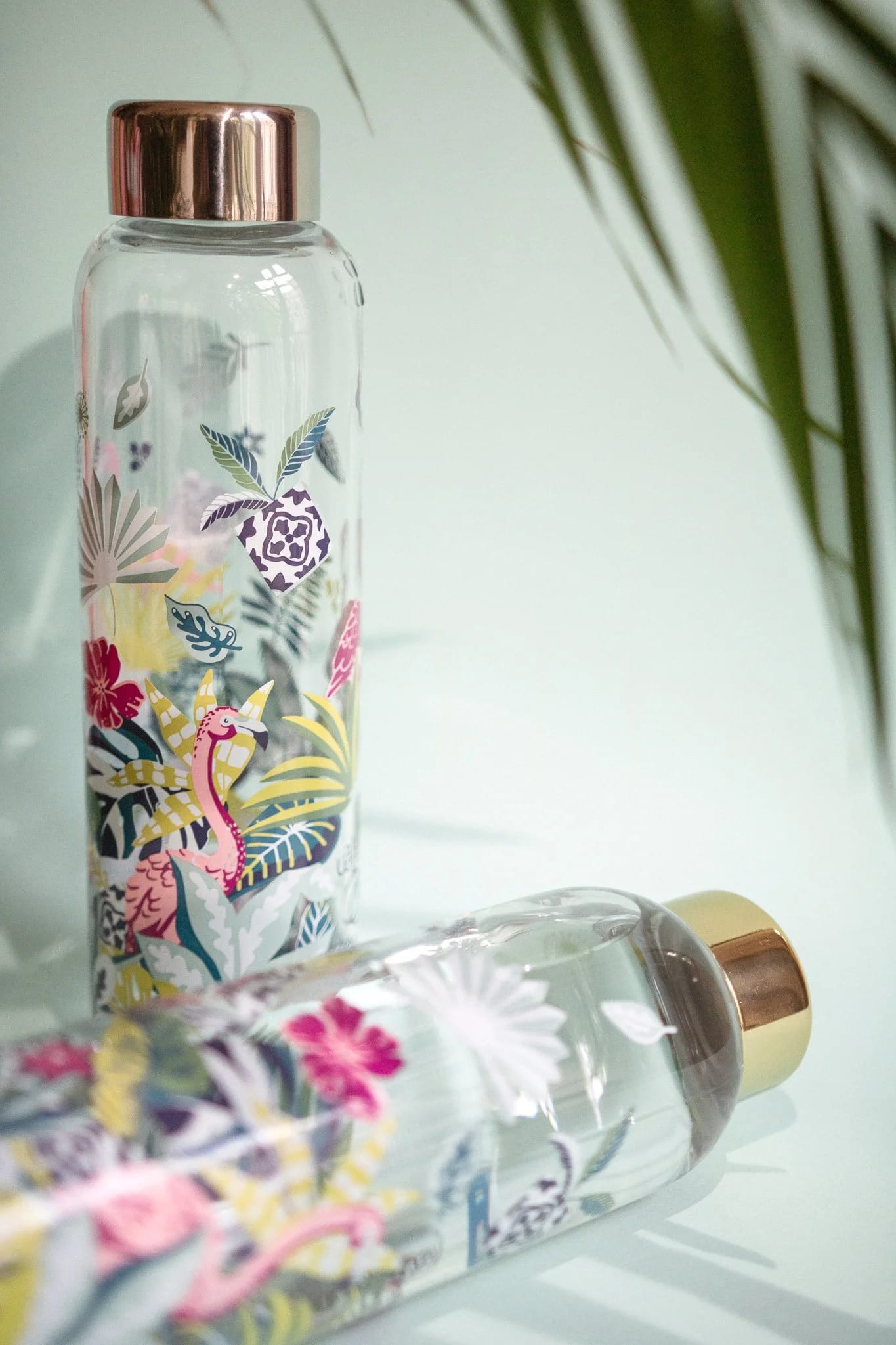 Two transparent glass water bottles with colorful tropical floral and dinosaur illustrations, one standing upright with a pink metallic cap and the other lying on its side with a gold metallic cap. Green leaves partially visible in the top right corner.